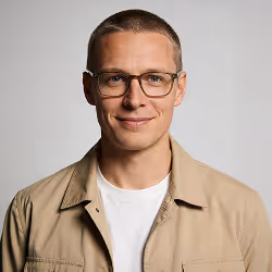 Smiling man with glasses wearing a beige jacket and white shirt against a gray background.