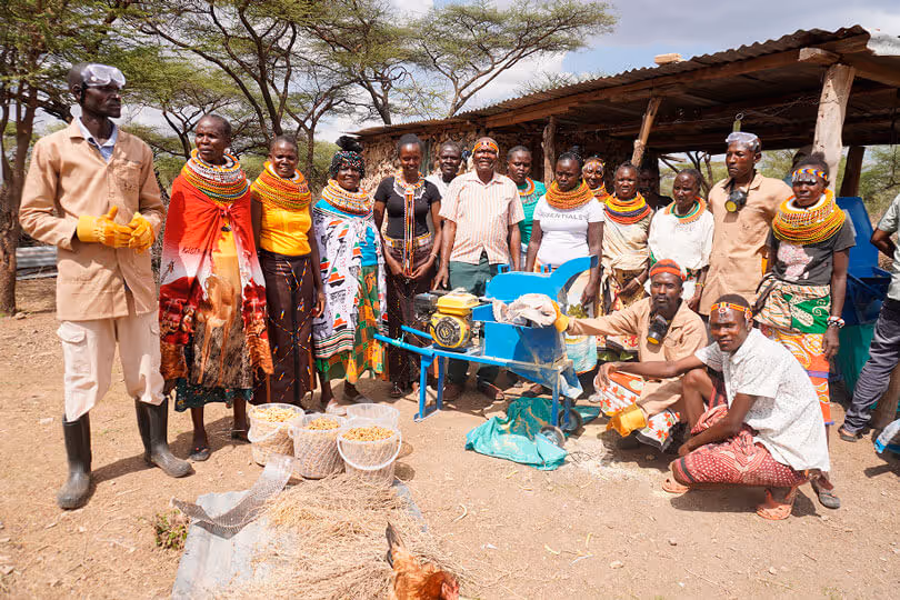 Gruppe afrikanischer Dorfbewohner in bunter Kleidung neben blauer landwirtschaftlicher Maschine im Freien.