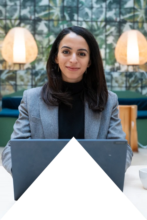 Jeune femme souriante aux cheveux noirs travaillant sur un ordinateur portable dans un espace de travail moderne avec des lampes et un mur décoré de plantes en arrière-plan.