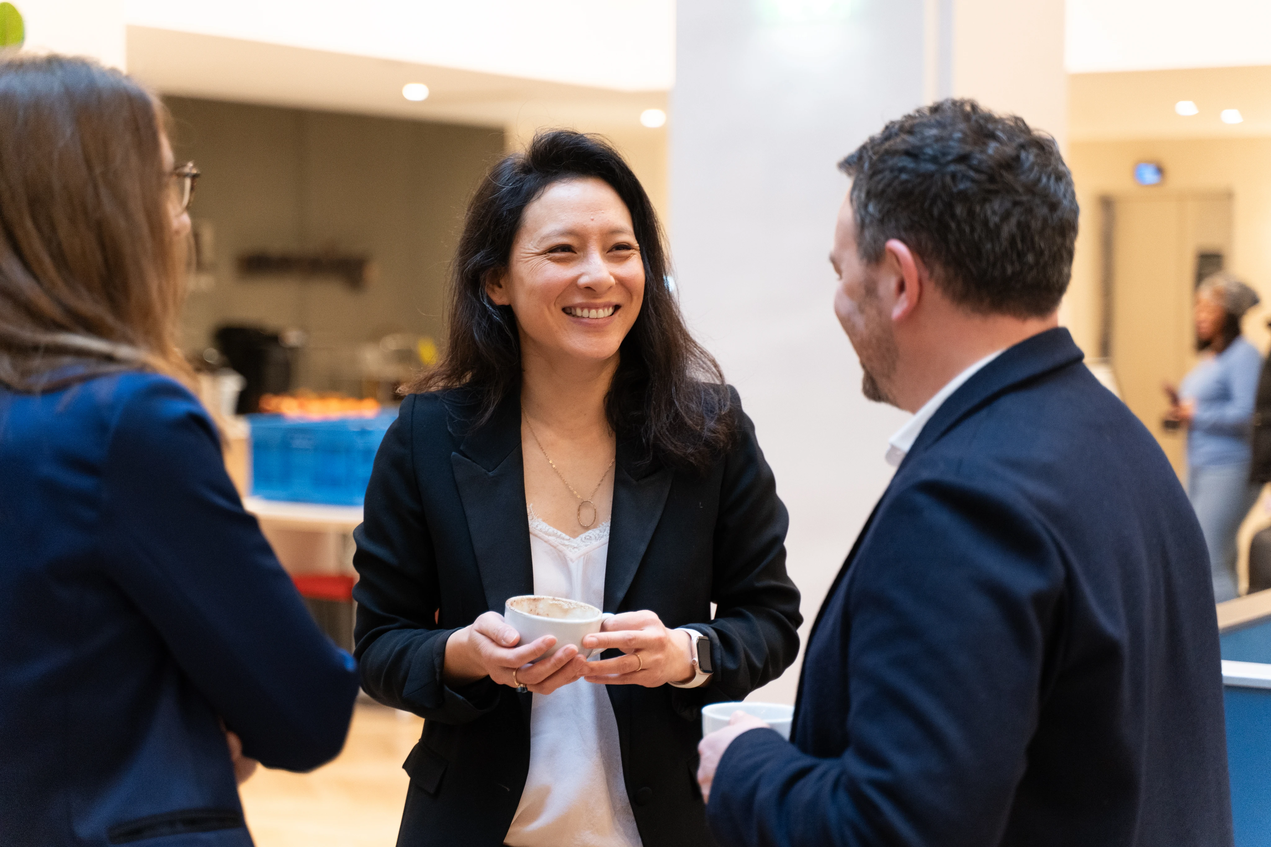 Trois personnes en tenue professionnelle discutent joyeusement, tenant des tasses de café dans un environnement de bureau lumineux.