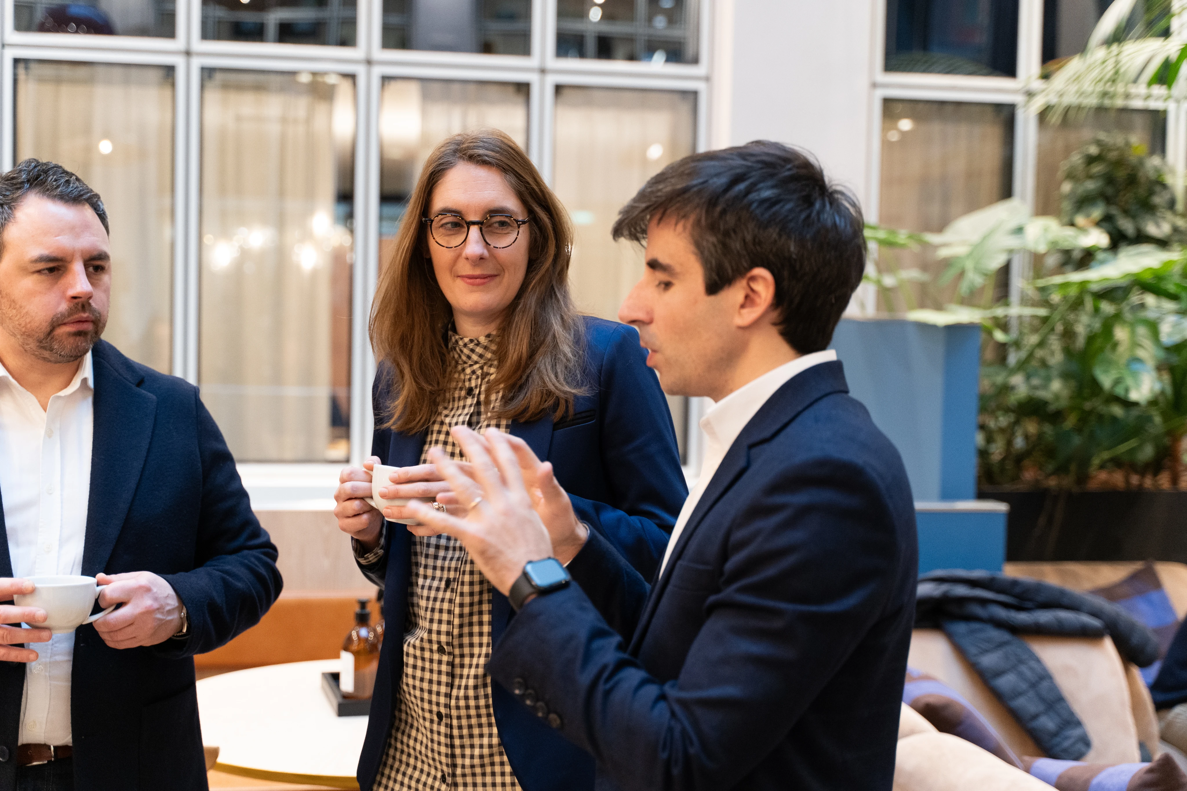 Trois professionnels discutant lors d'une pause café dans un espace intérieur lumineux avec des plantes.