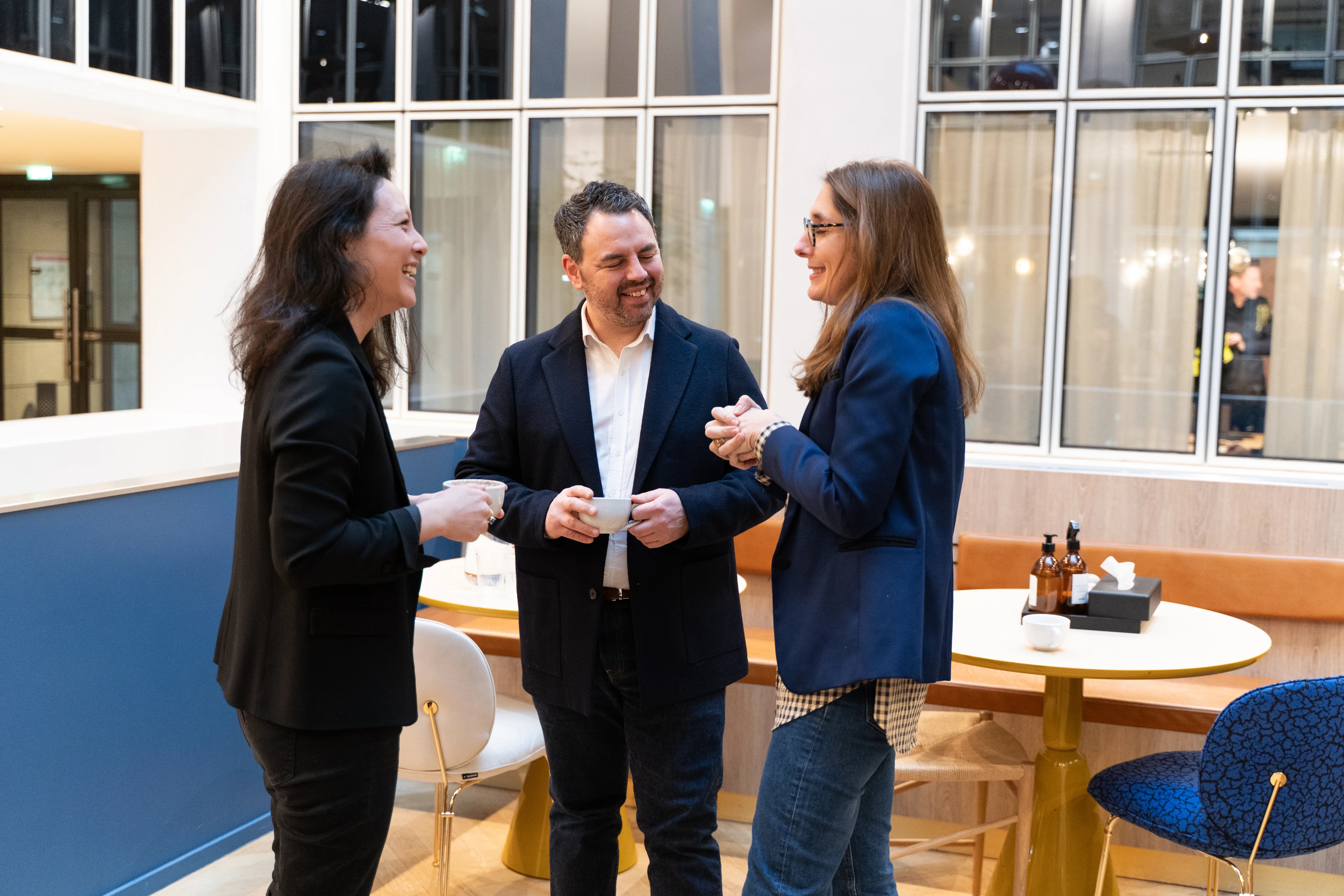 Trois personnes souriantes discutant dans un espace de bureau moderne avec des tasses à la main.