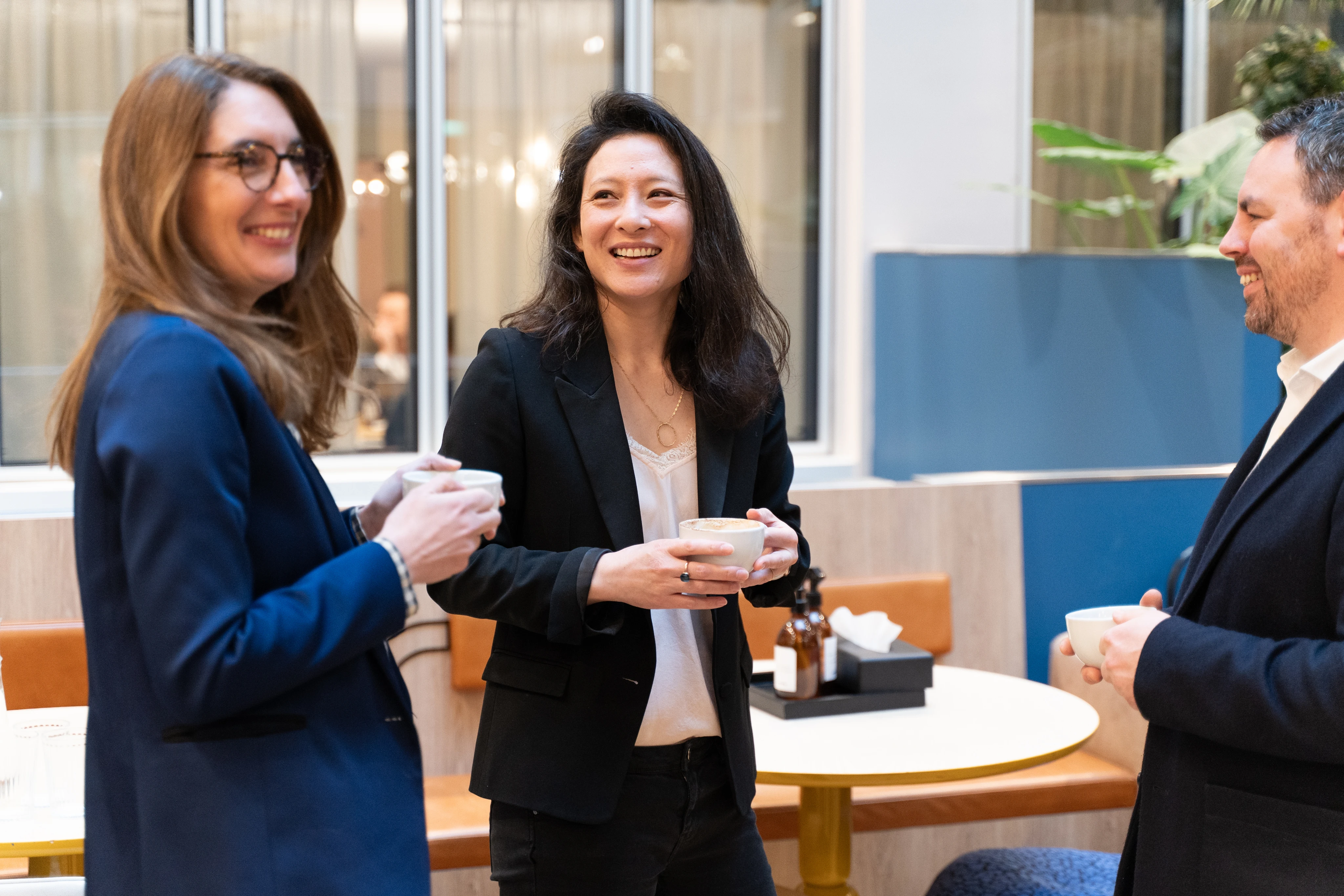 Trois collègues souriants tenant des tasses de café lors d'une conversation dans un espace de bureau moderne.