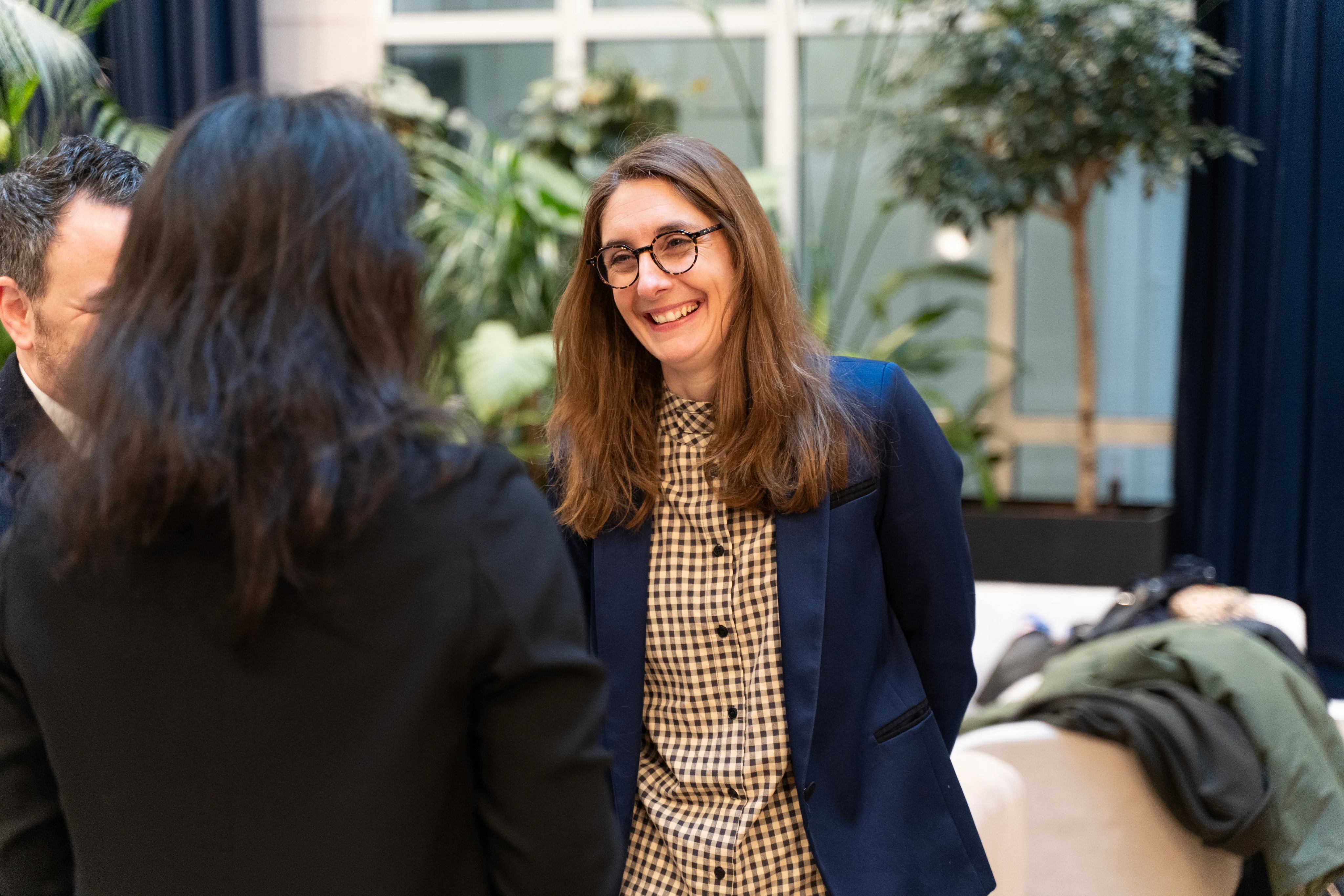 Une femme souriante portant des lunettes, une veste bleue et une chemise à carreaux discute avec deux autres personnes dans un espace intérieur avec des plantes.
