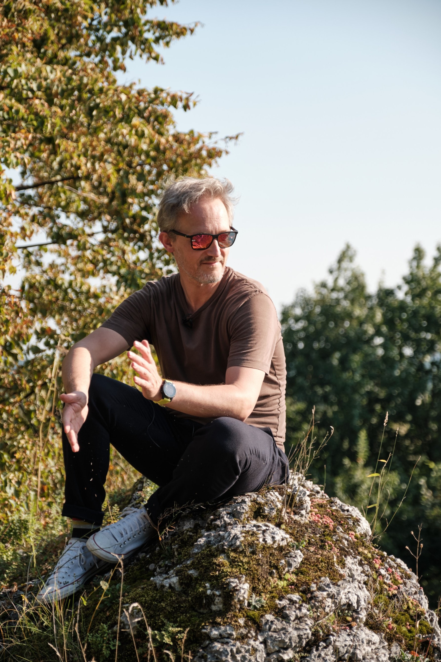 Man wearing sunglasses sitting on a moss-covered rock outdoors with trees in the background.