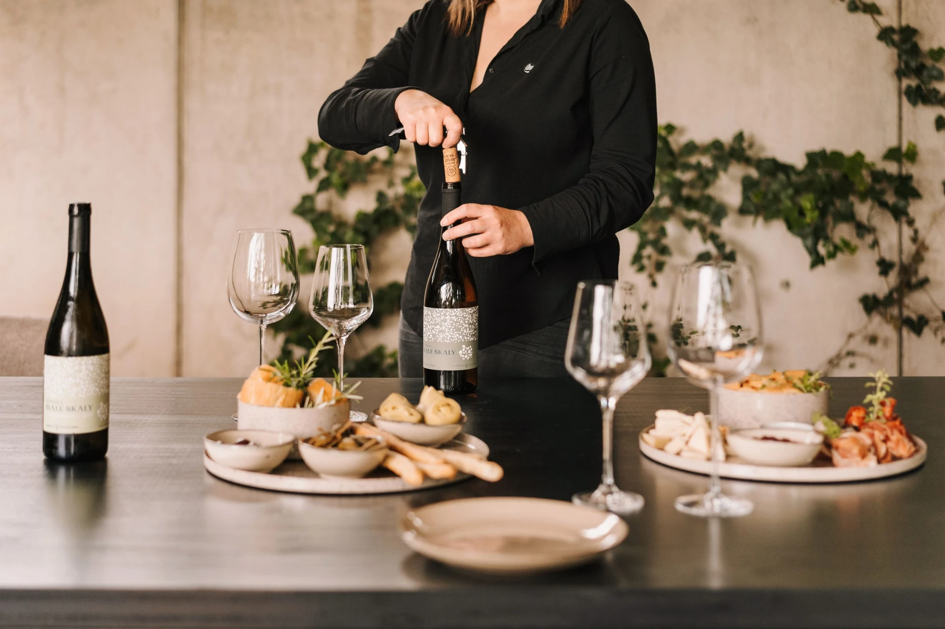 Person opening a bottle of wine at a table set with wine glasses, plates, and a variety of appetizers.