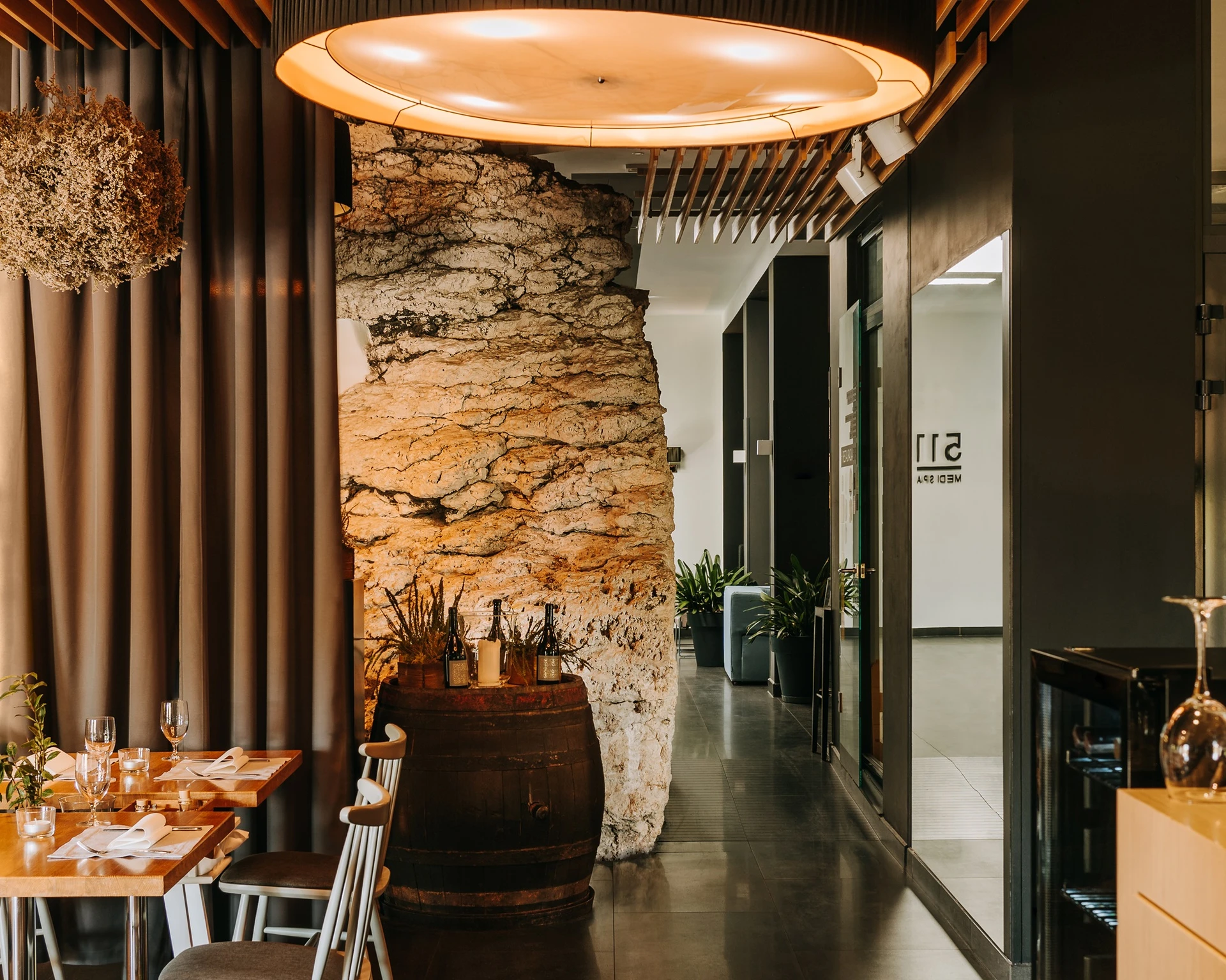 Cozy restaurant interior with wooden tables, chairs, a large barrel with bottles, and warm lighting.