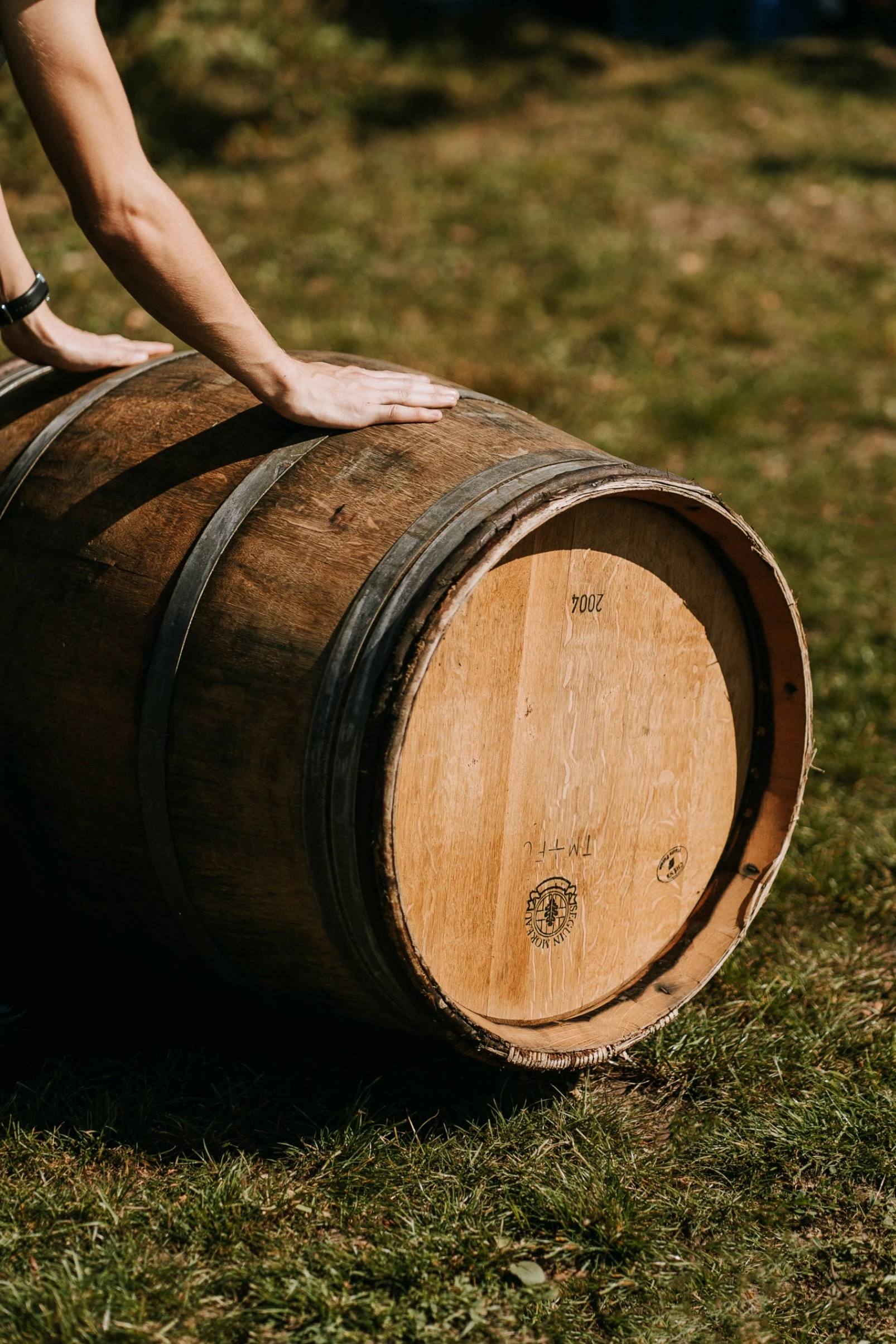 Person's hands resting on a wooden barrel lying on grass in sunlight.