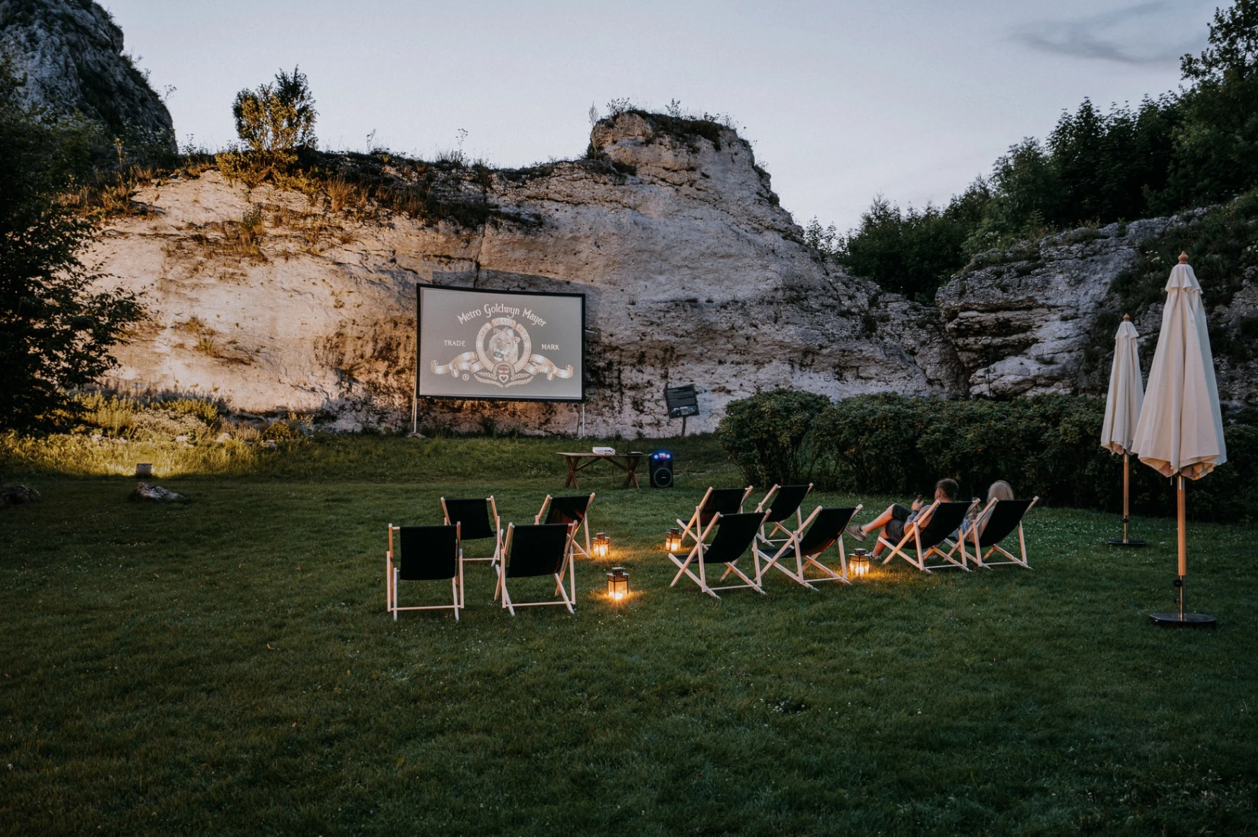 Outdoor movie setup in a grassy area with deck chairs facing a screen against a rocky cliff during dusk.