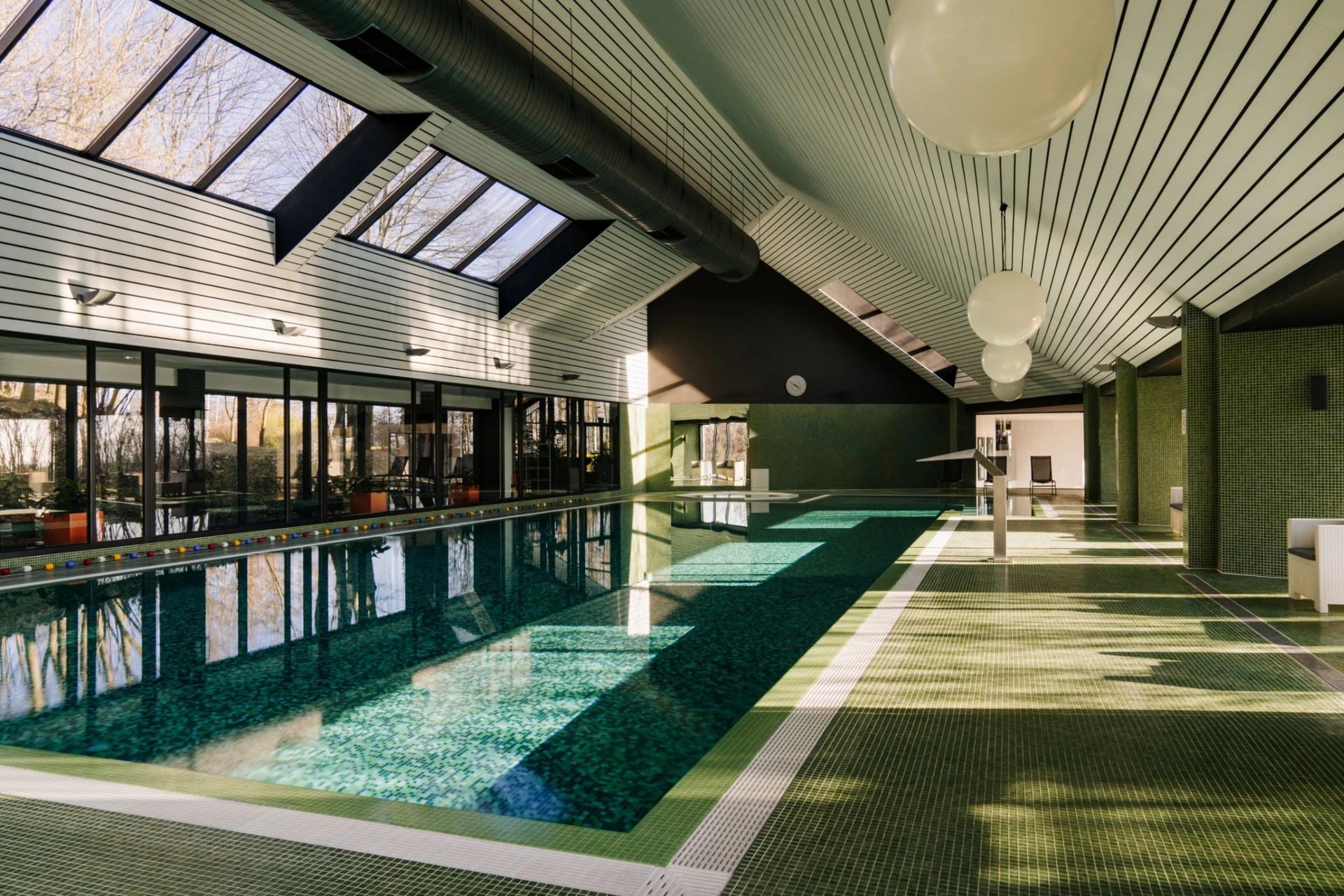 Indoor swimming pool with green mosaic tiles, large windows, skylights, and round hanging lights.
