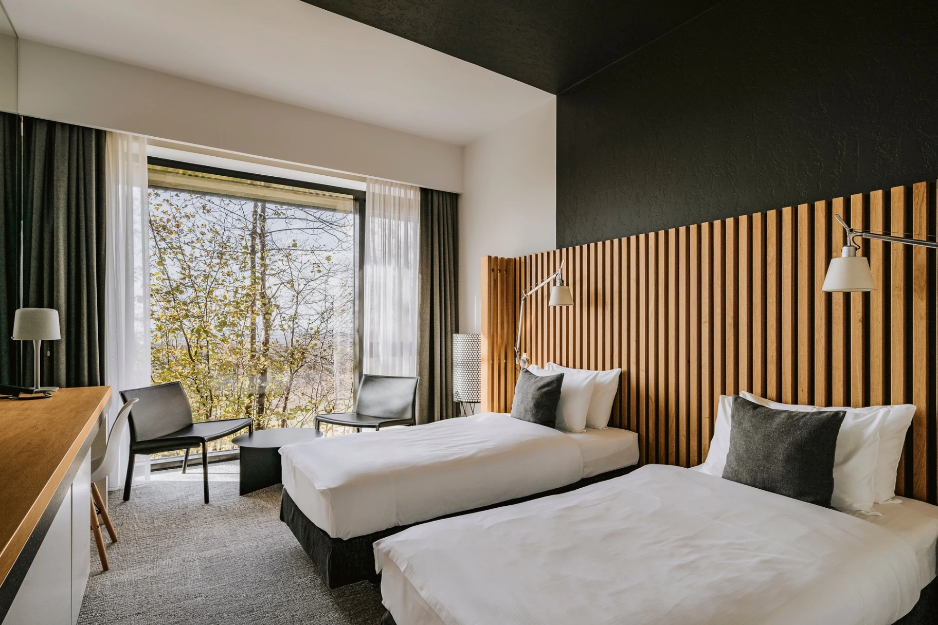 Modern hotel room with two single beds, wooden slat headboard, black and white pillows, and large window with a view of trees.