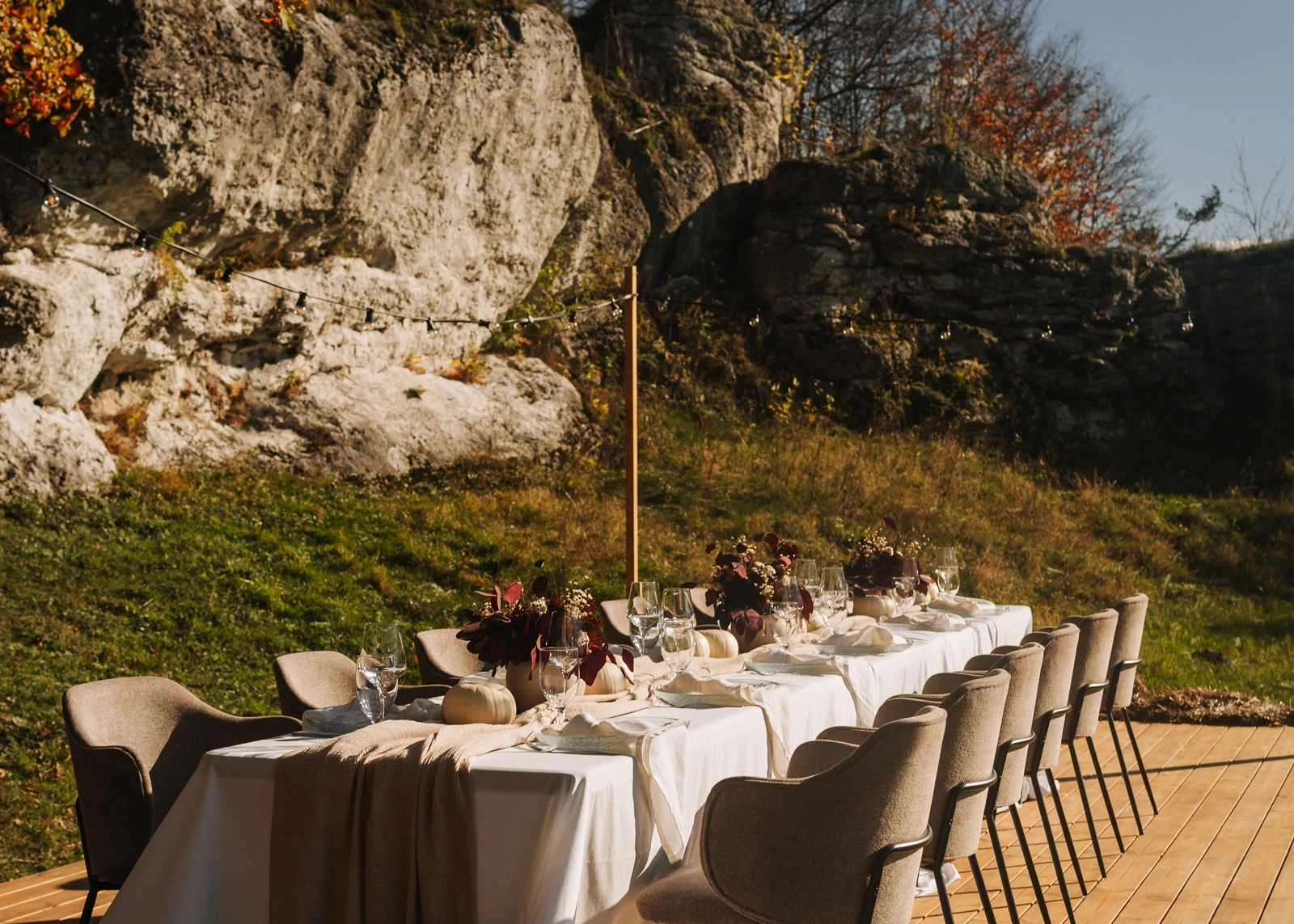 Outdoor dining table set with white tablecloth, fall-themed floral arrangements, white pumpkins, and glassware on wooden deck near rocky hillside.