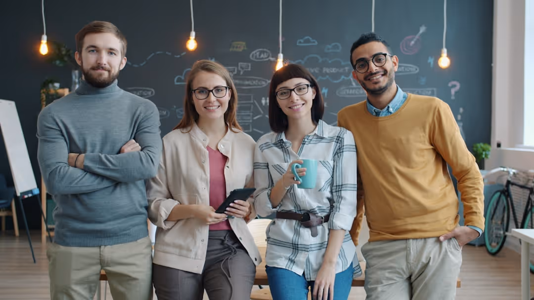 Four young professionals standing in a modern office, smiling, with a chalkboard wall and hanging light bulbs in the background.