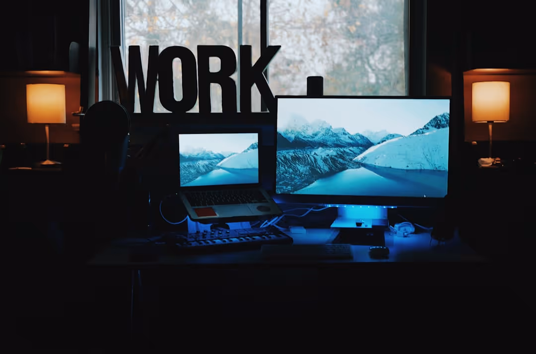 Dark workspace with two screens showing mountain landscapes, illuminated by two lamps and blue LED light under the desk.