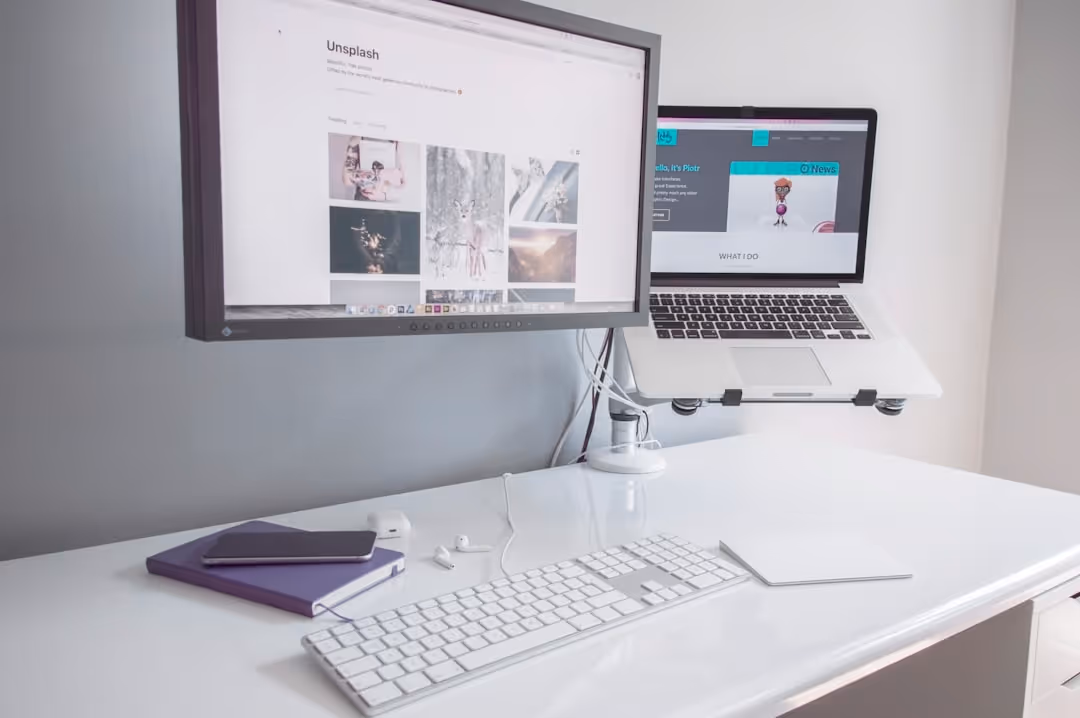 Clean white desk with a wireless keyboard, trackpad, smartphone on a notebook, earbuds, and dual monitors, one a desktop screen and the other a laptop computer mounted on an adjustable arm.