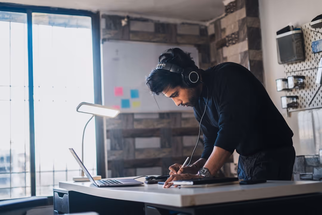 Man wearing headphones, leaning over a desk while writing on a notepad next to an open laptop and desk lamp in a well-lit room.