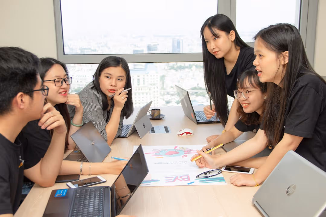 A group of six young adults seated around a table in a modern office, engaged in a discussion with laptops and a colorful infographic on the table.