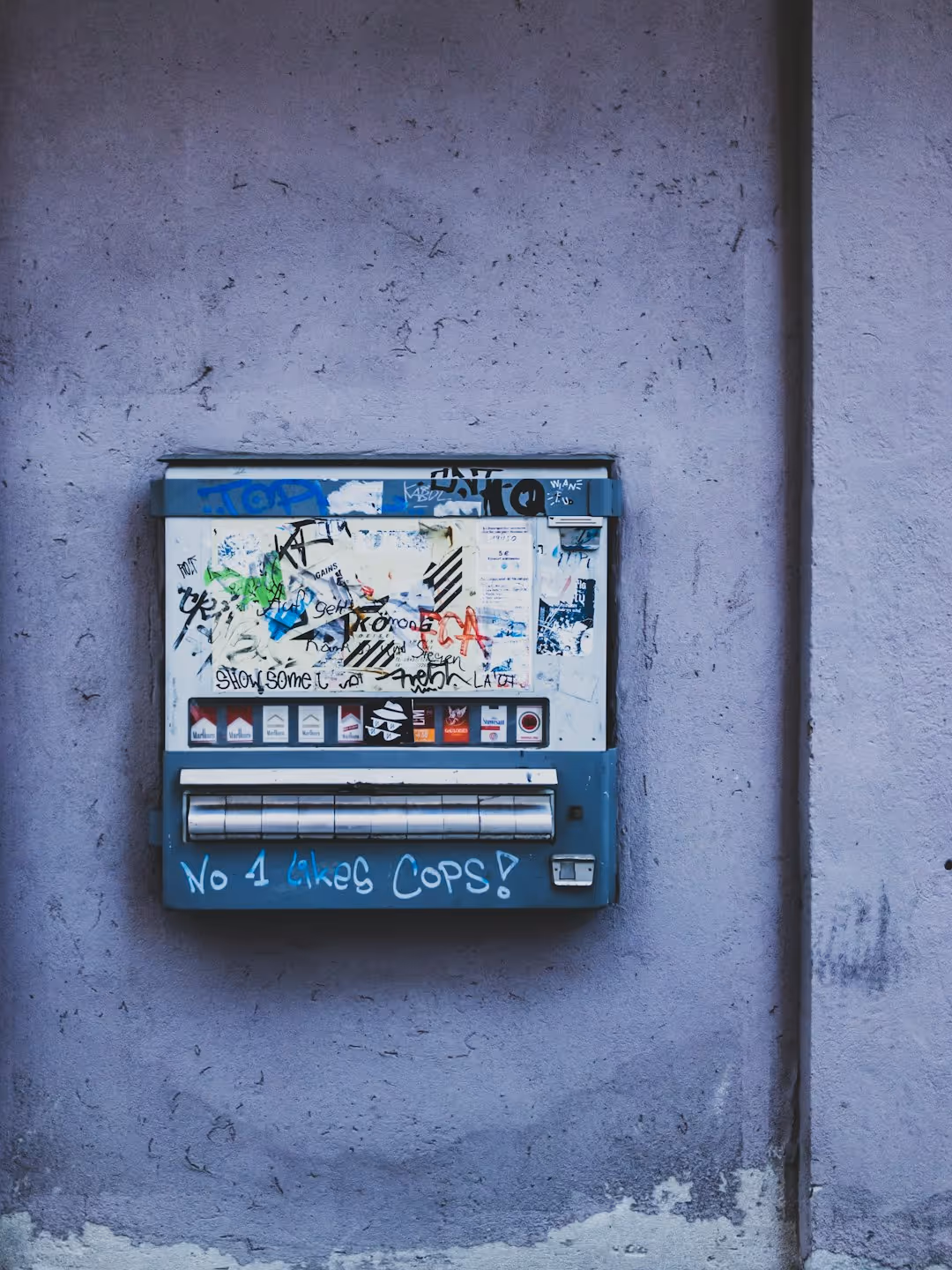 Old cigarette vending machine with graffiti and stickers, mounted on a worn purple wall.