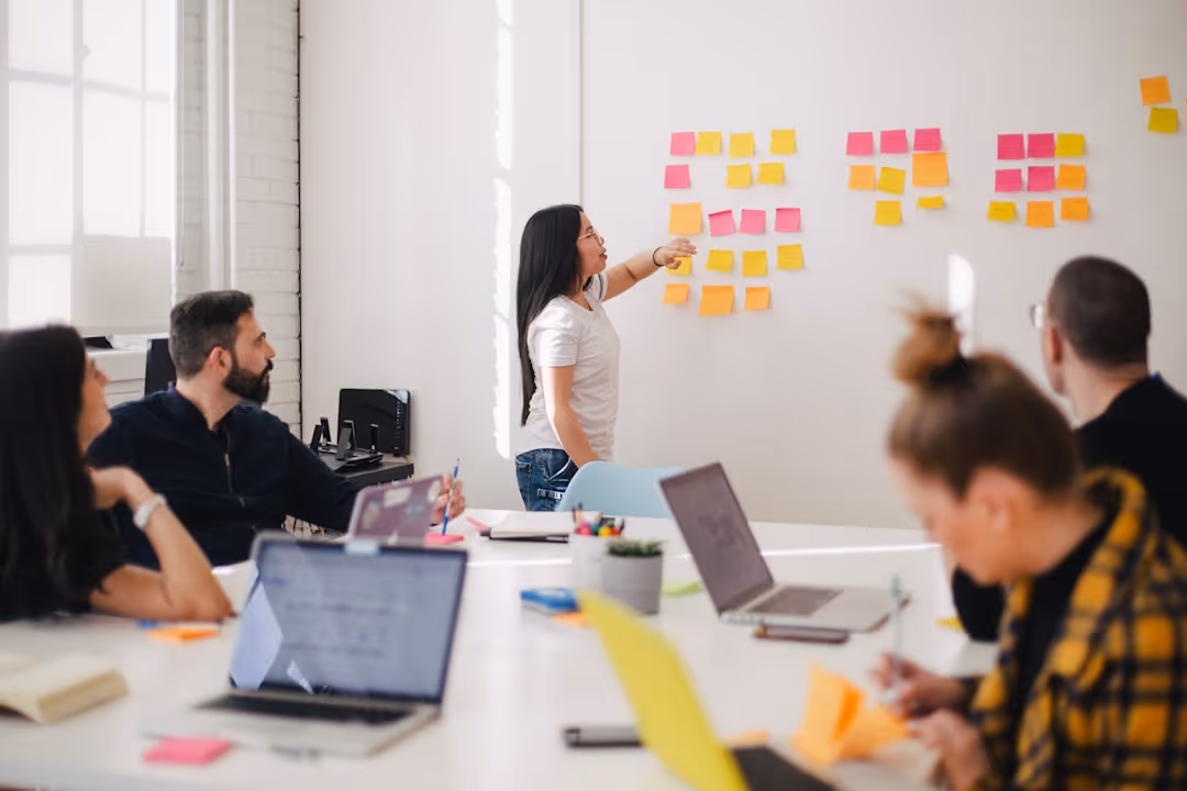 A woman presenting ideas on sticky notes on a whiteboard while colleagues listen and work on laptops in a bright meeting room.