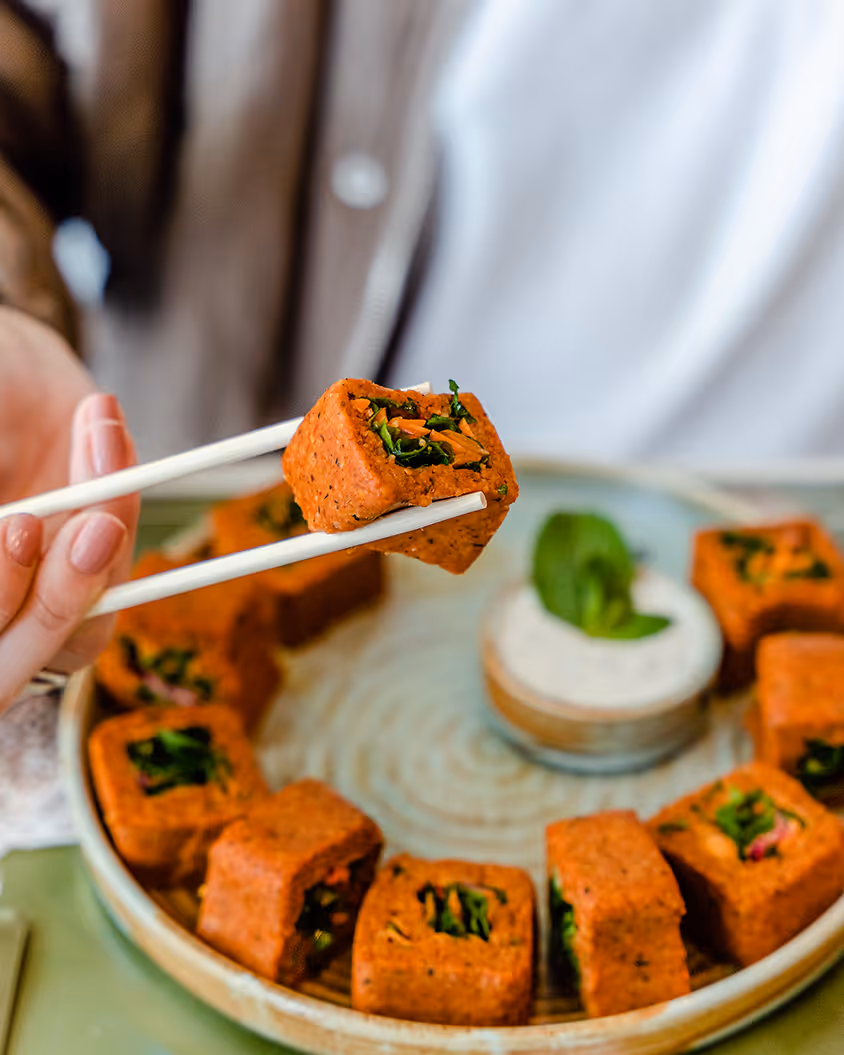 Hand hält mit Essstäbchen ein gewürztes rotes veganes Gericht, umgeben von weiteren gleichen Stücken auf einem Teller mit einer kleinen Dipschale.