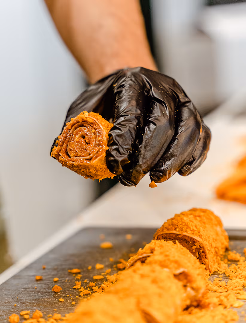 Hand in schwarzem Einweghandschuh hält ein Stück eines mit Keksbröseln bedeckten Schokoladenröllchens über einer Schneidefläche mit weiteren Stücken.