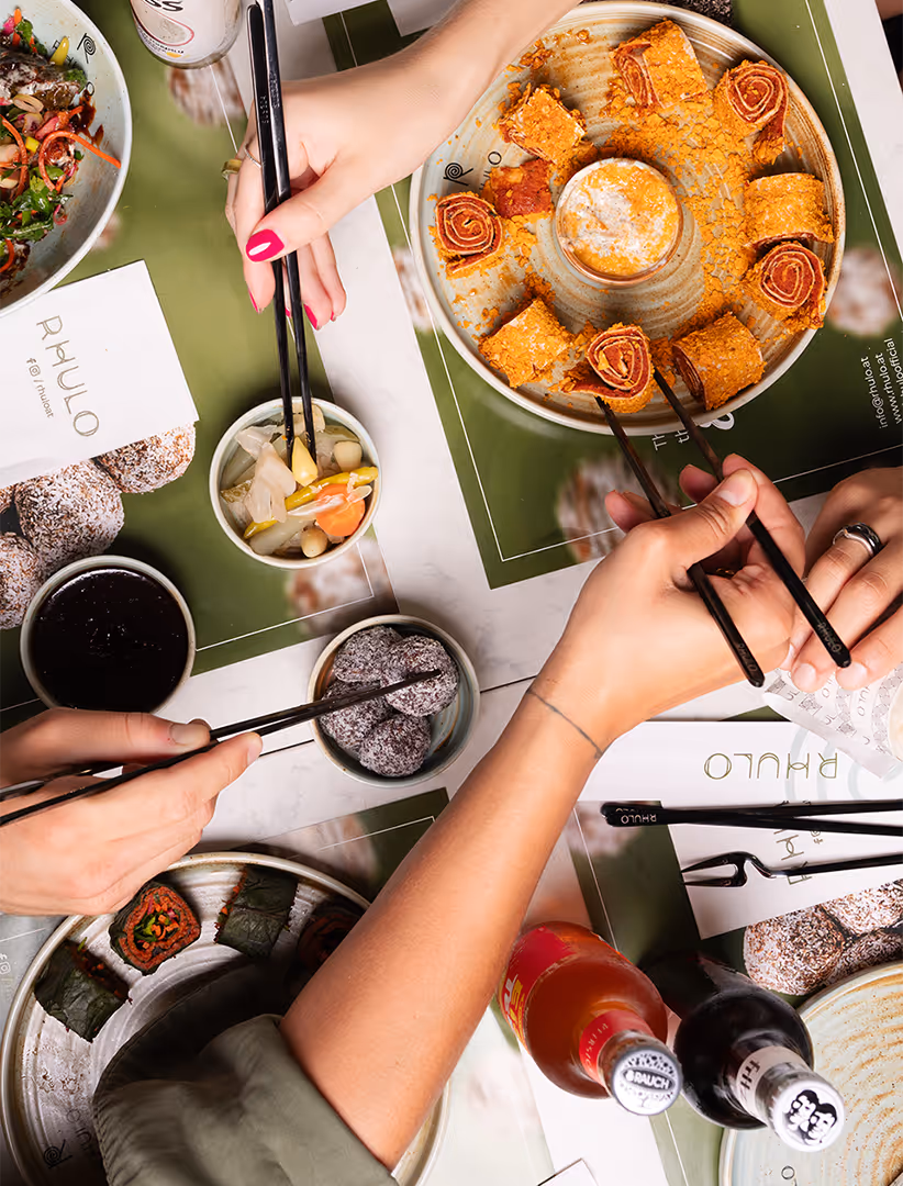 Mehrere Hände mit Essstäbchen greifen gemeinsam koreanisches Essen von Tellern auf einem Tisch mit grün-weißem Rhulo-Design.