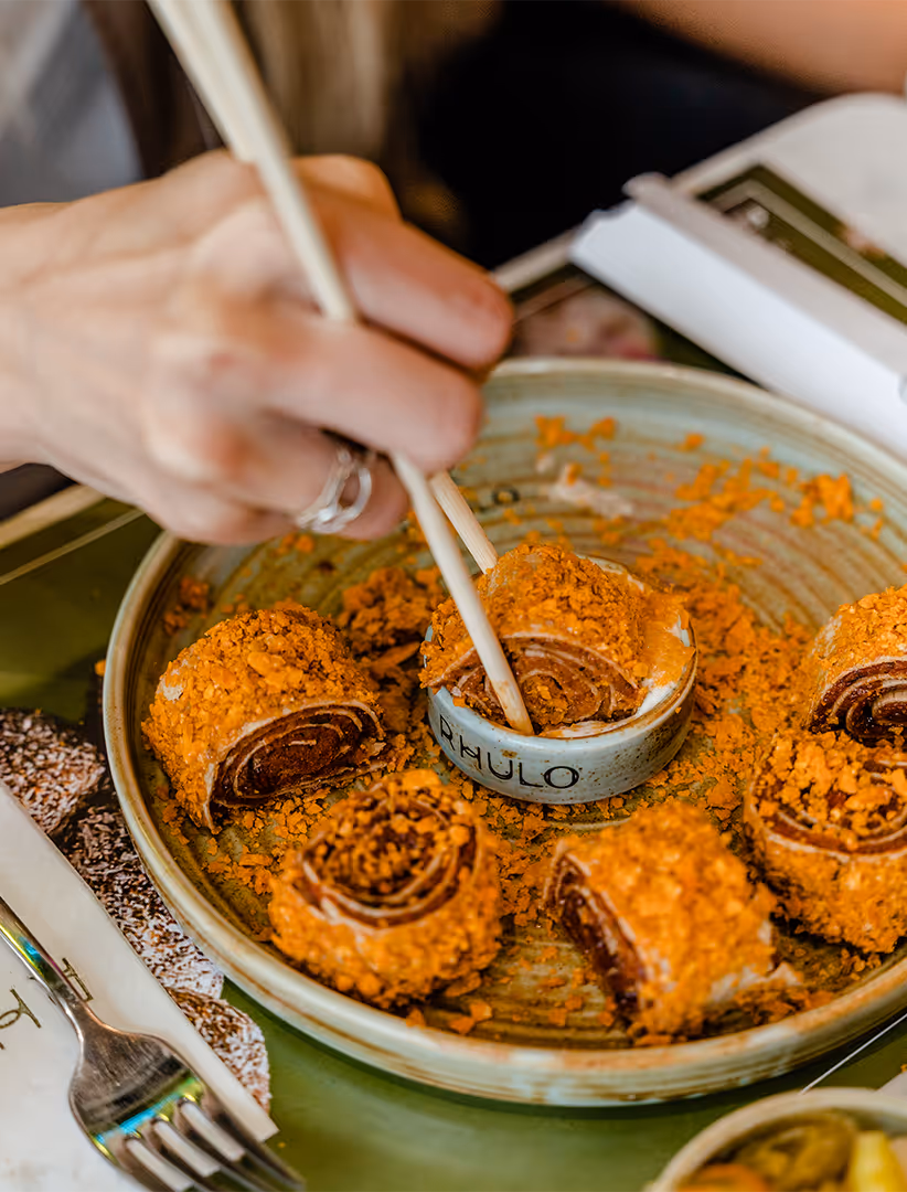 Hand hält Stäbchen und taucht ein mit knusprigem, orange-bestreutem Sushi-Röllchen in eine kleine Schale mit Soße.