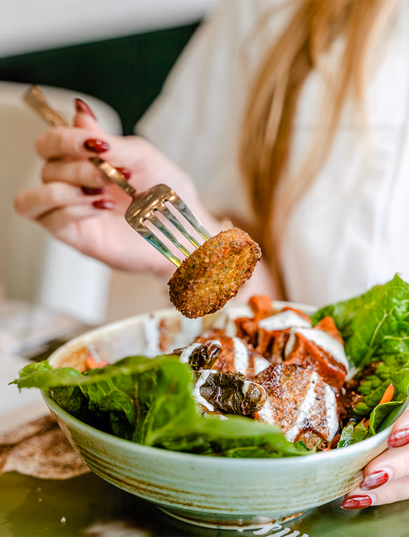 Hand hält eine Gabel mit einem Falafel über einer Schüssel mit Salat und Joghurt-Dressing.