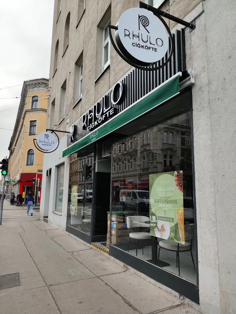 Außenansicht des Rhulo Çiğköfte Ladens mit einem grünen Banner und Schaufenstern auf einem städtischen Bürgersteig.