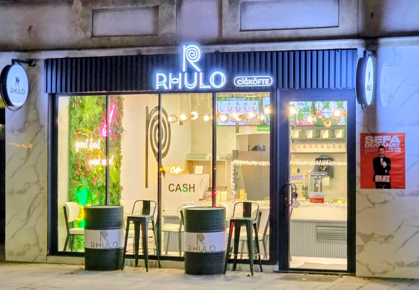 Beleuchteter Eingangsbereich eines Restaurants namens Rhulo Çiğköfte mit Sitzgelegenheiten und grüner Pflanzenwand.