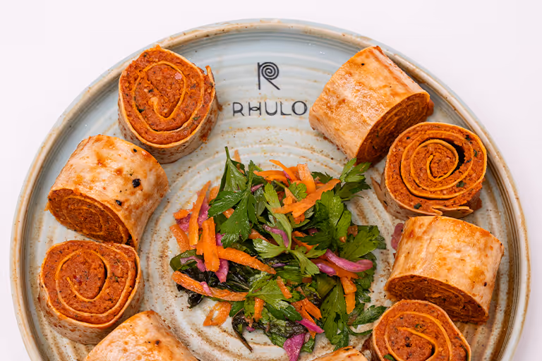 Teller mit sechs aufgerollten Wrap-Scheiben und einem Beilagensalat aus Karotten, Zwiebeln und Petersilie in der Mitte.