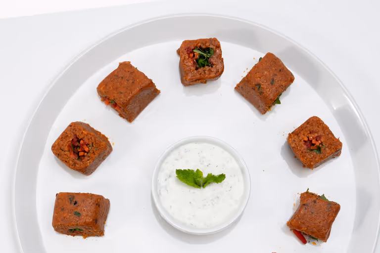 Weiße Platte mit acht rechteckigen veganen Köfte, angeordnet in einem Kreis um eine Schale mit Joghurt-Dip, garniert mit Minzblatt.