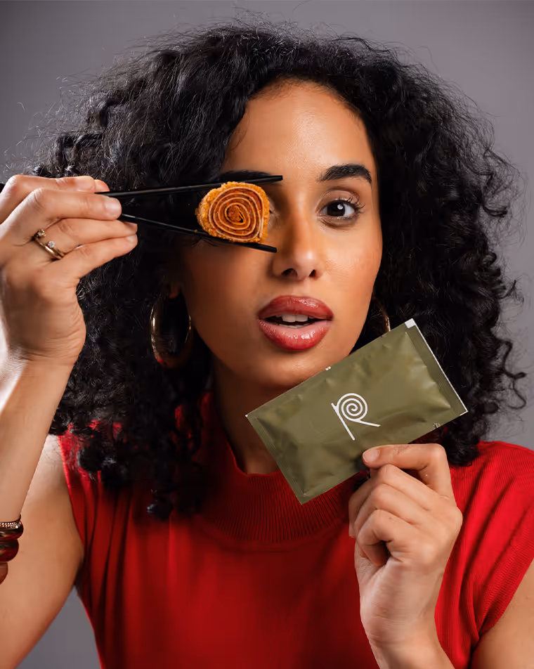 Frau mit lockigem Haar in rotem Oberteil hält mit Essstäbchen eine aufgerollte Sushi-ähnliche Speise vor ihr Auge und in der anderen Hand eine grüne Verpackung mit stilisiertem spiralförmigen Logo.