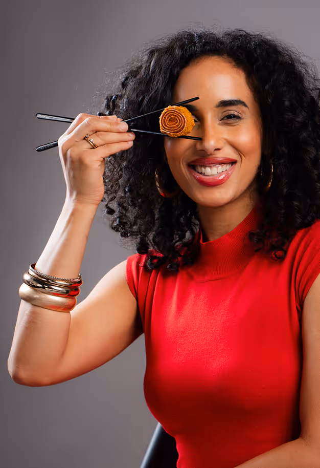 Frau mit lockigem Haar und rotem Top hält ein Sushi-Stück mit Essstäbchen vor ihr Auge und lächelt.