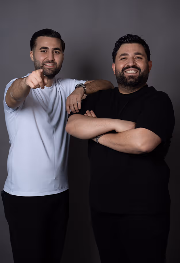 Zwei Männer mit Bart lächeln vor grauem Hintergrund; einer trägt weißes T-Shirt und zeigt nach vorne, der andere trägt schwarzes T-Shirt und hat die Arme verschränkt.