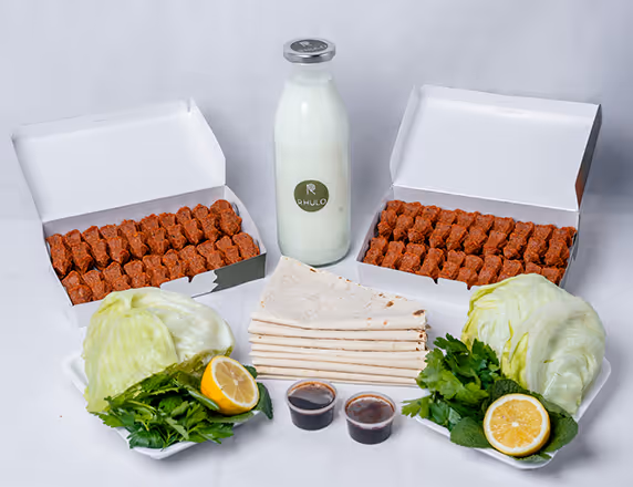 Zwei geöffnete Schachteln mit traditionellen türkischen Çiğ Köfte, Fladenbrot, zwei Eisbergsalathälften, Zitronen, frischen Kräutern, zwei kleinen Behältern mit Soßen und eine Flasche Milch.