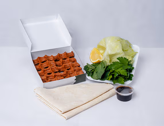 Kästchen mit Çiğ Köfte, ein Paket Fladenbrot, frische Salatblätter, Kräuter, Zitronenschnitz und Soße auf weißem Hintergrund.