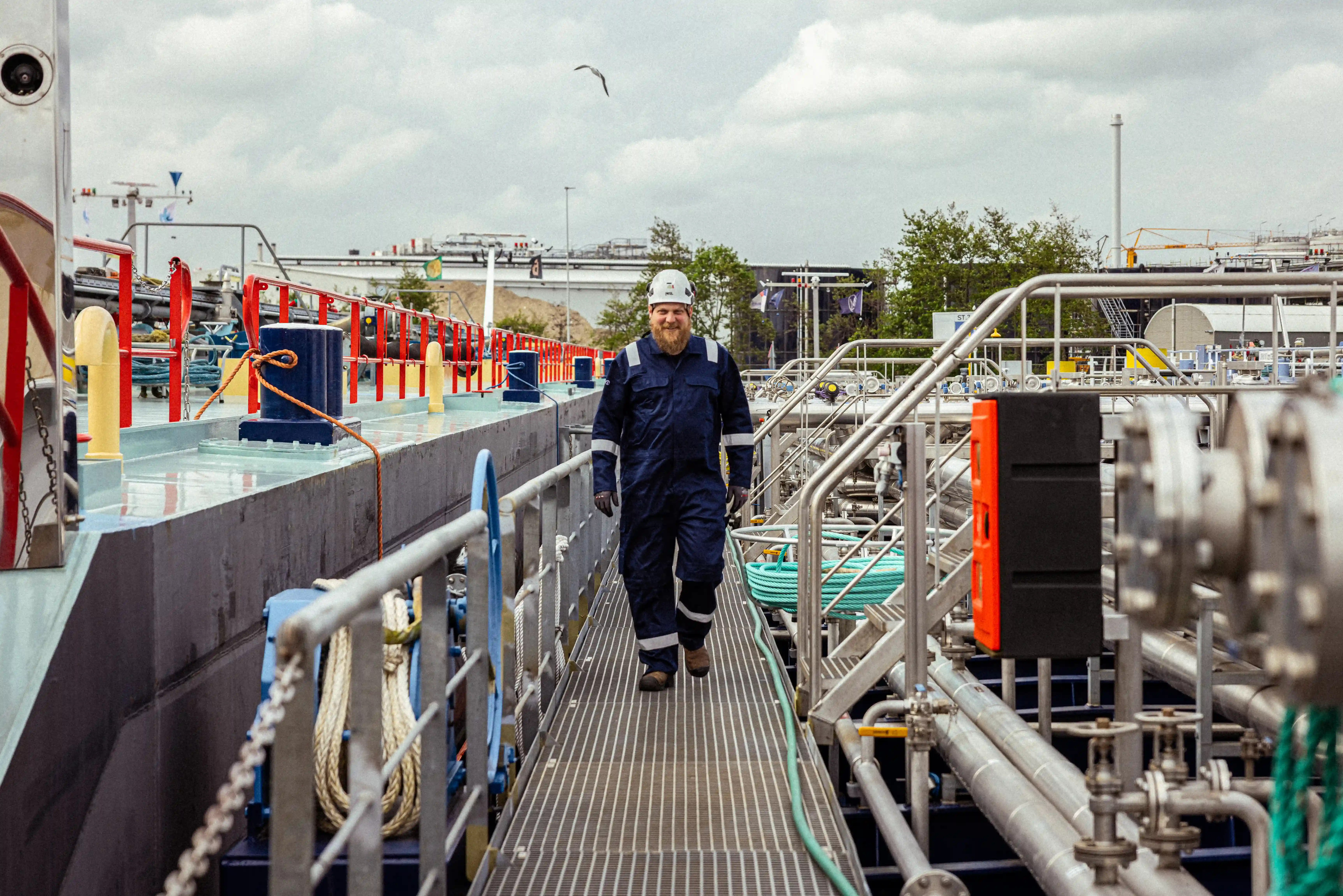 Een medewerker van Mercurius, gekleed in een blauwe overall met een witte helm, op het dek van een binnenschip in een industriële haven.