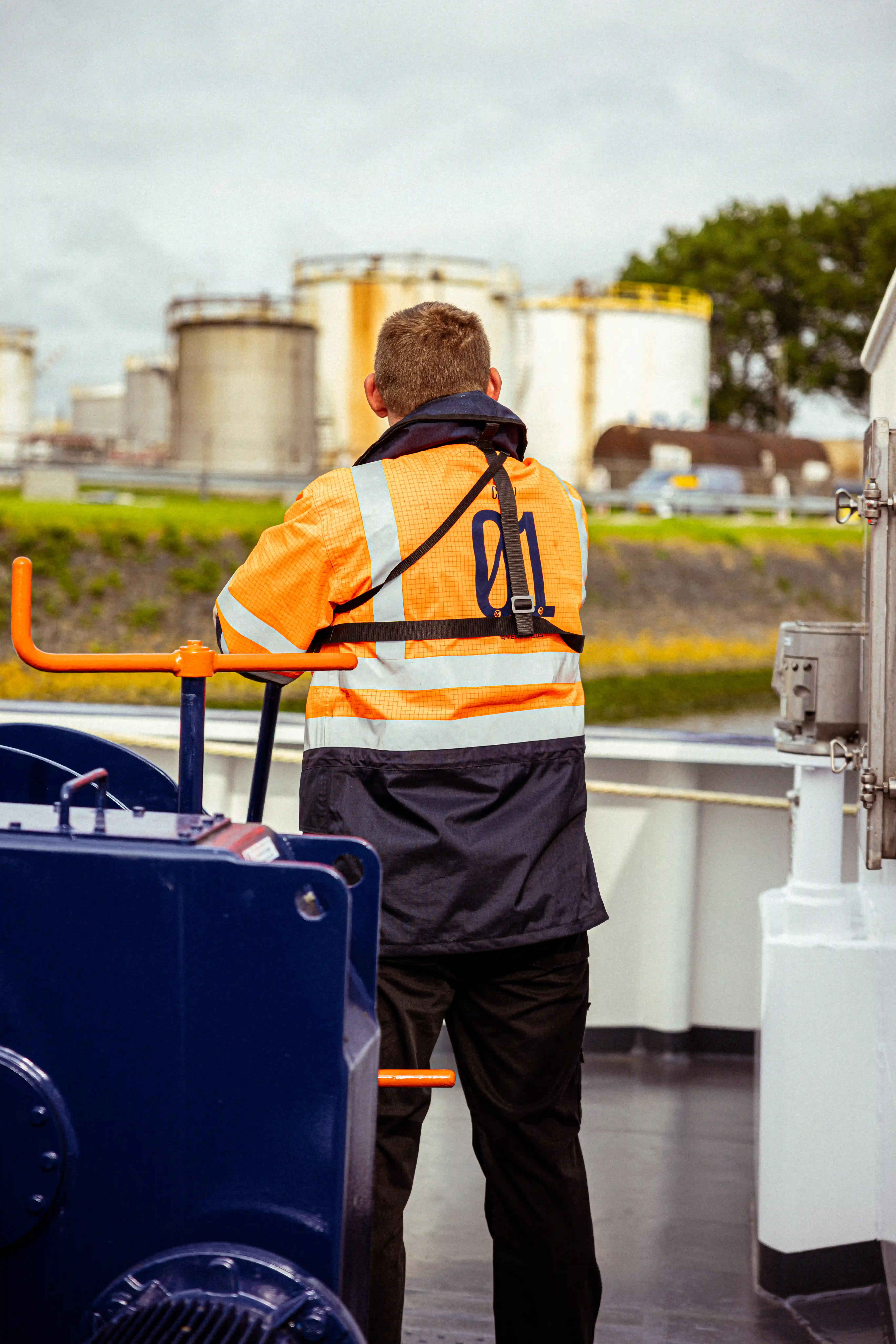 Een medewerker van Mercurius, gekleed in een oranje werkjas met 'CREW 01' op de rug, op het dek van een binnenschip in een industriële haven.