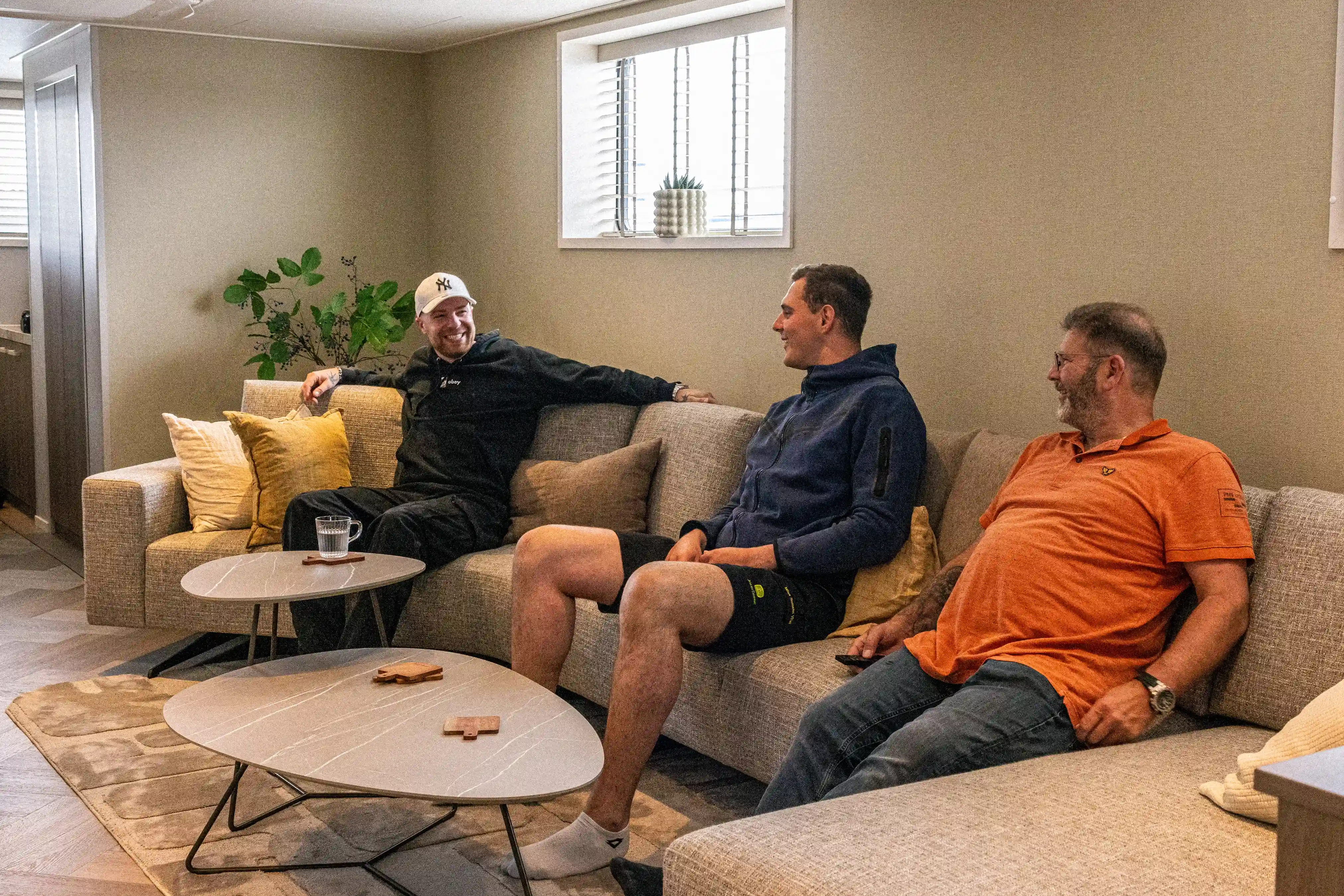 Drie bemanningsleden van Mercurius ontspannen en in een lachend gesprek op een moderne beige bank in de leefruimte van een binnenschip