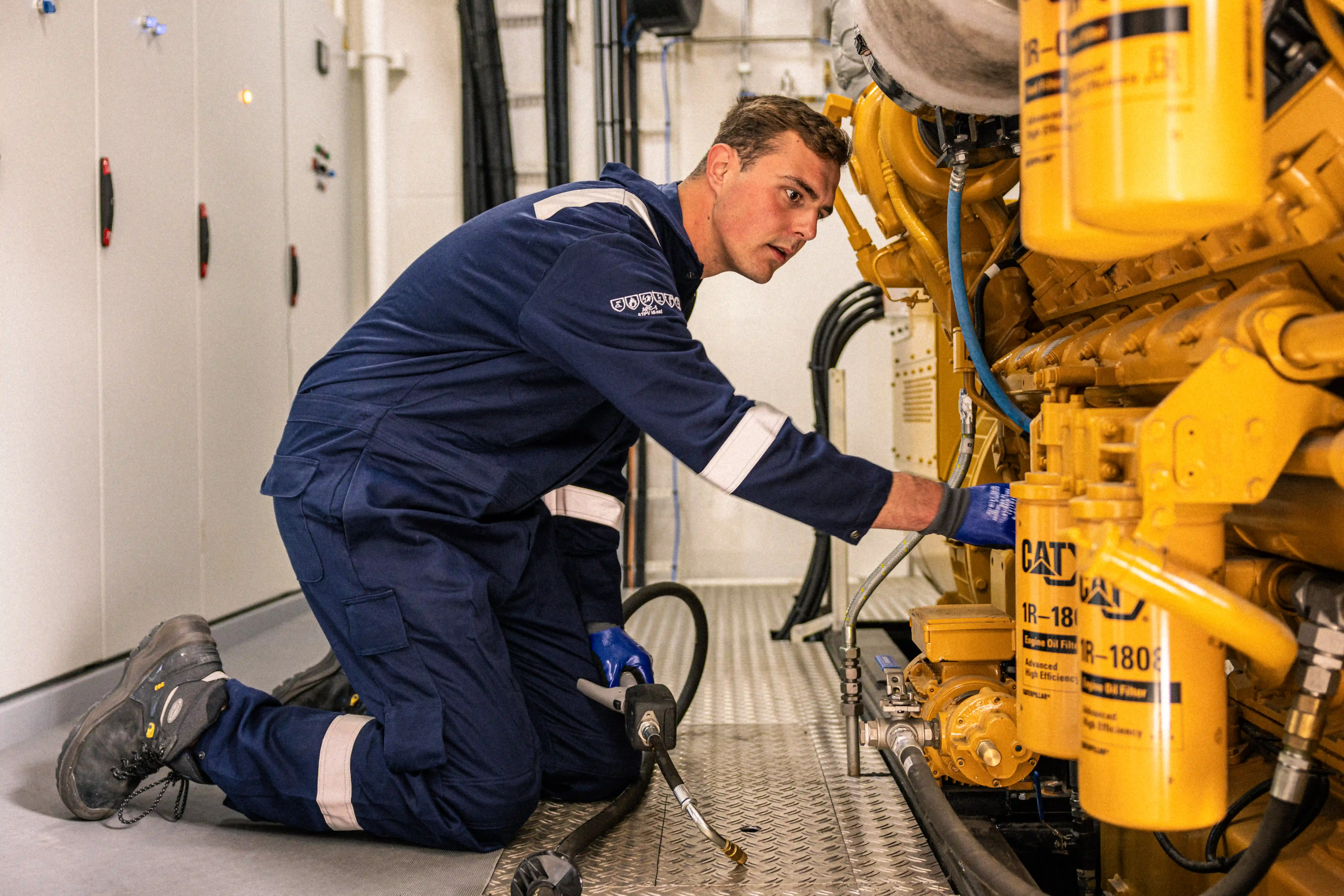 Bemanningslid in blauwe overall werkt aan een grote gele Caterpillar motor in de machinekamer van een schip