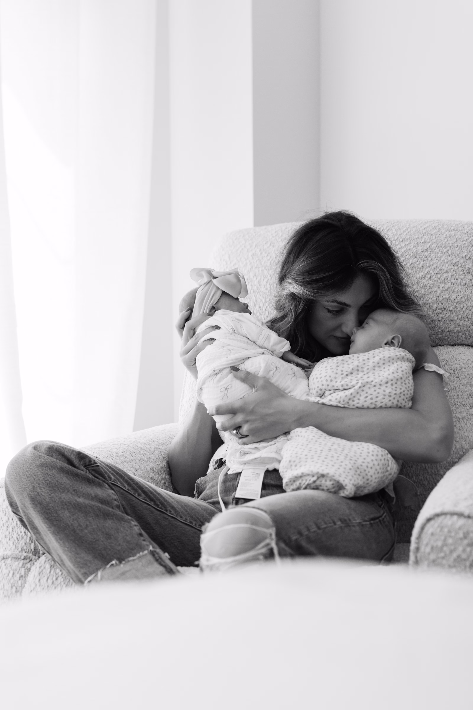 Mother sitting in a chair lovingly holding and touching noses with her two swaddled newborn babies.