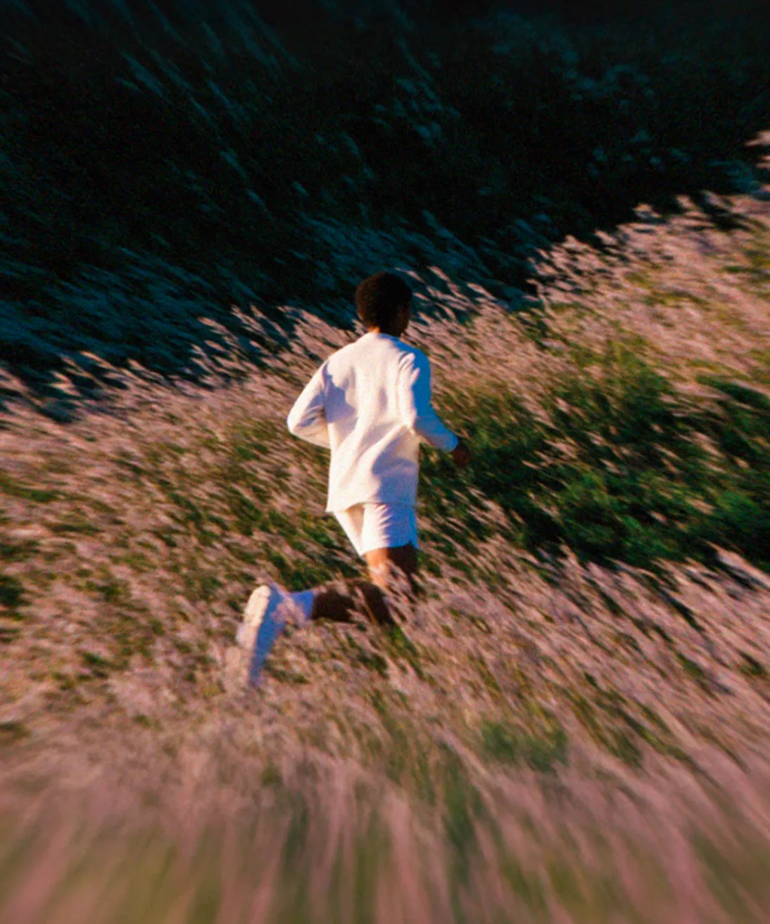 Person in white outfit running through a field of tall grass or wildflowers during sunset or dusk.