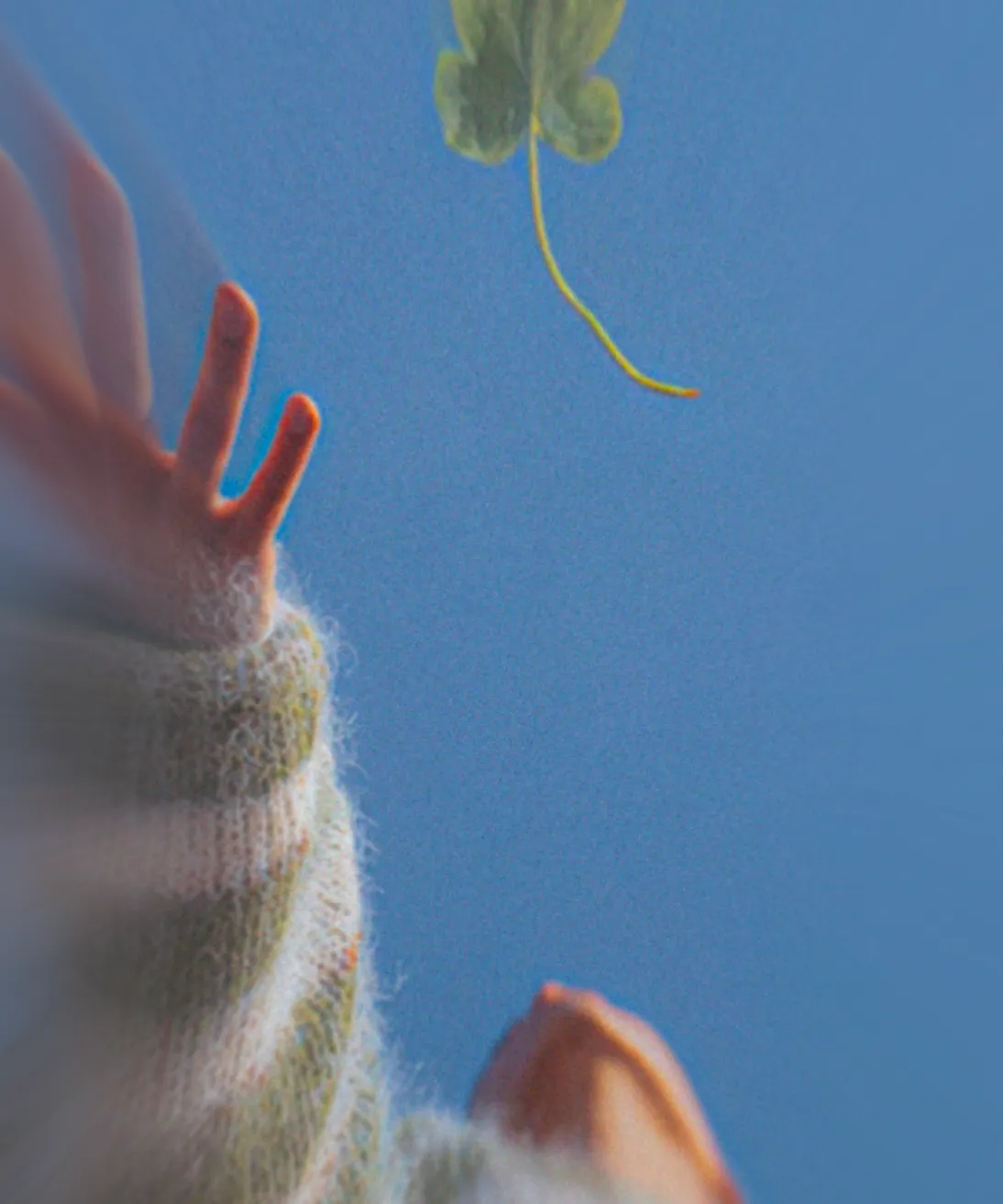 Person reaching up towards a four-leaf clover against a clear blue sky.