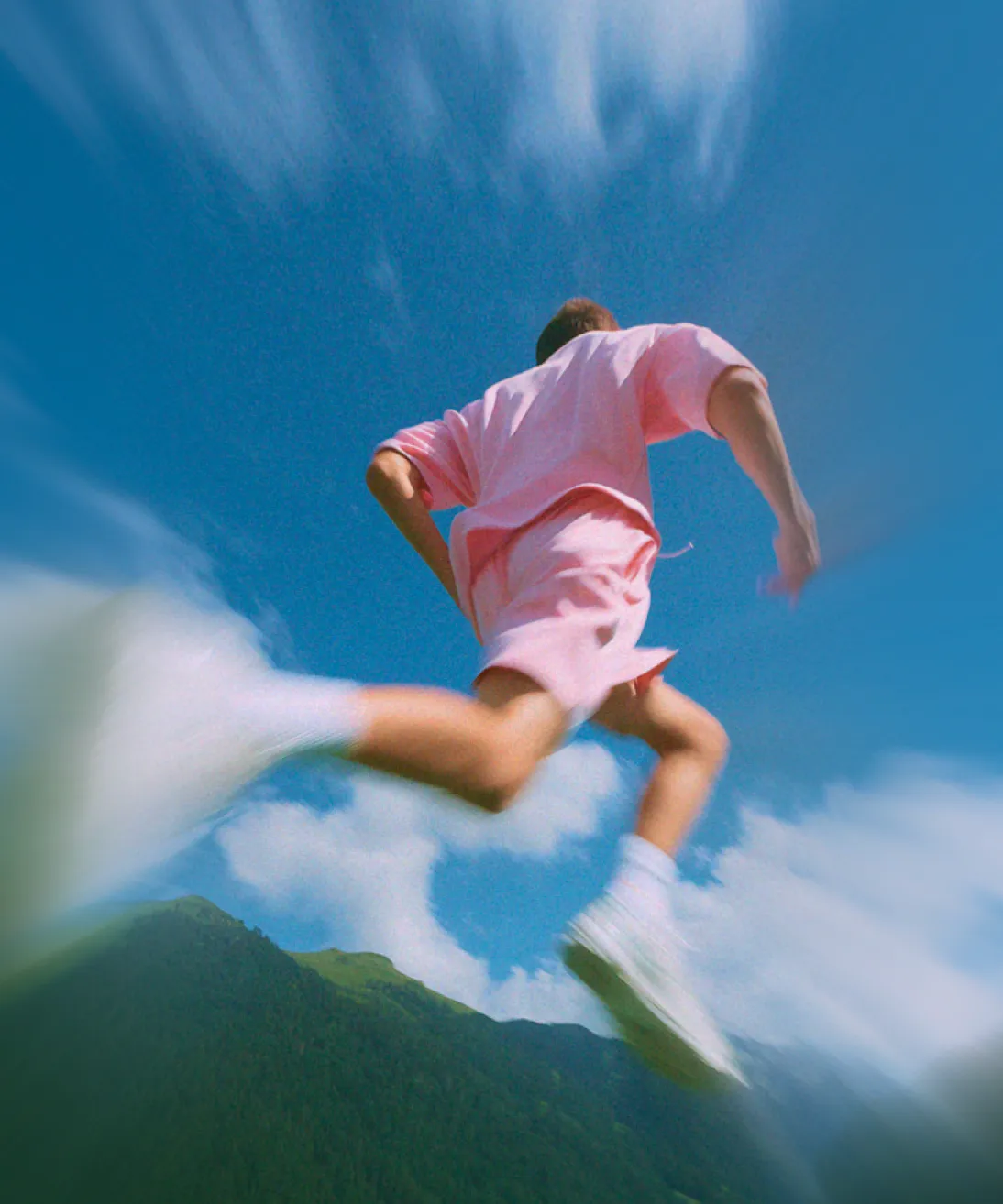 Person in pink clothes running outdoors against a blue sky and green mountain background.