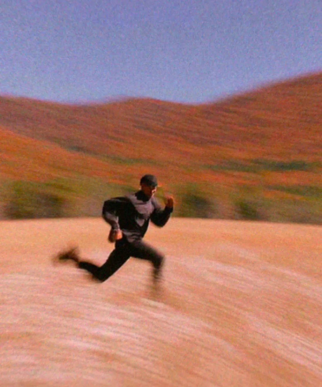 Person in black running rapidly across a dry, open landscape with hills in the background.