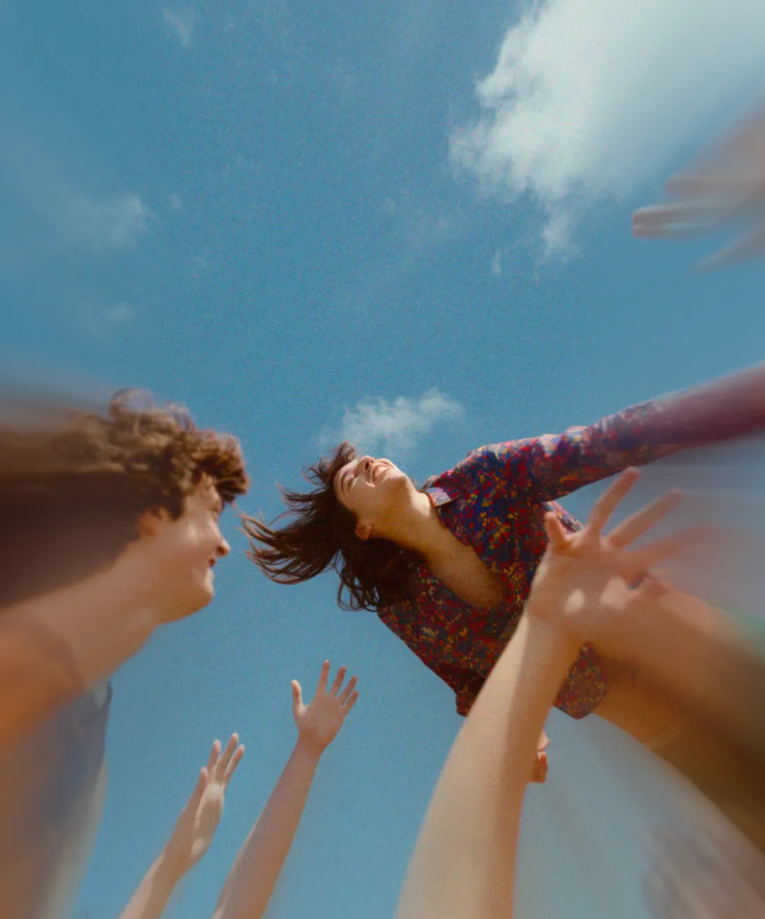 Young woman with dark hair laughing while being lifted into the air by friends under a blue sky with clouds.