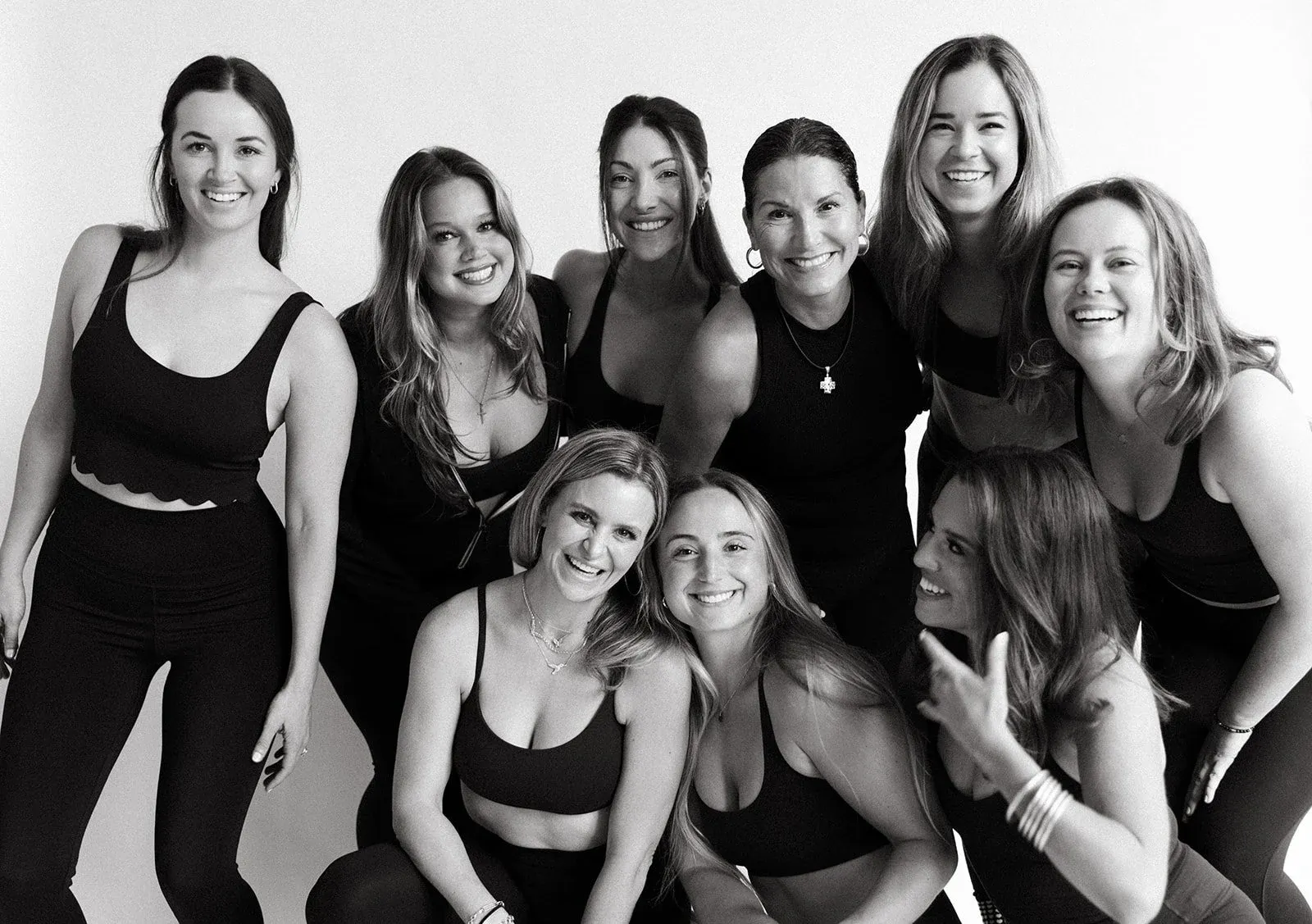 Group of nine smiling women posing together in black athletic wear against a plain background.