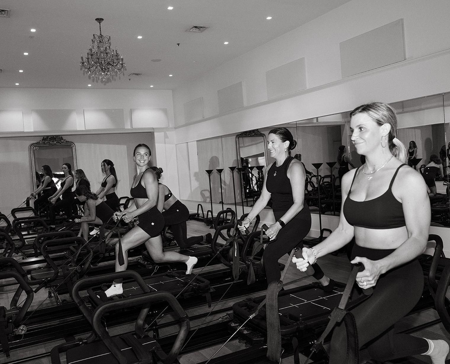 Women exercising on Pilates reformer machines in a bright studio with mirrors and chandeliers.