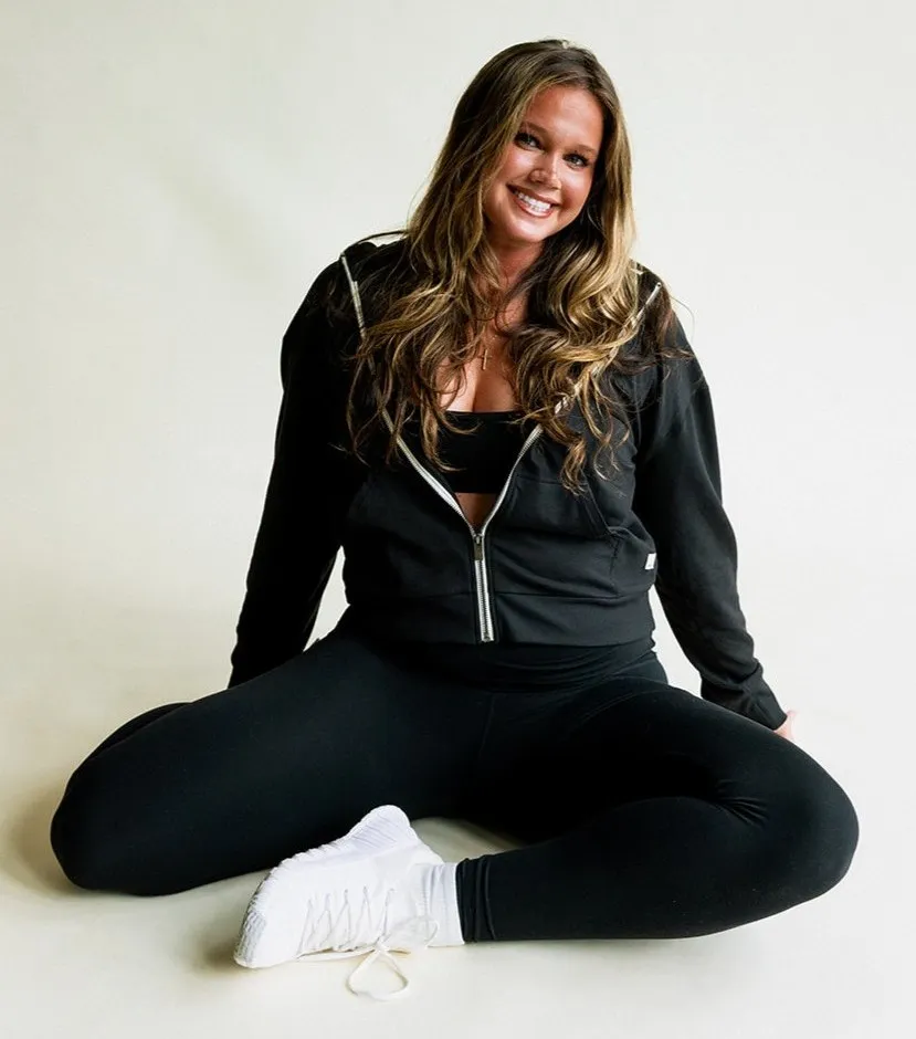 Smiling woman in black activewear sitting on the floor with legs spread and white sneakers.